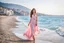 Placeholder: long shot, beautiful Turkish girl with nice blue eyes wearing a pink - blue dress walks in shore toward camera , sharp focus all the scene, natural light, vertical composition, relaxed and natural, fresh and comfortable ,in seaside there are luxury beach hotels.