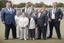 Placeholder: Overweight white politician wearing a suit; group of thin, muscular athletes in sports clothes; outside, sports field; realistic, photographic