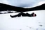 Placeholder: in the distance side a wiev a figure in dark clothes and long black leather coat lies on his back in the snow and looks the sky in a winter landscape, alone, white snow, high contrast, cold, winter, mountains, white, blue, gray and black colors, cinematic, atmospheric, dark, gloomy, best shot