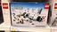 Placeholder: display of a box of the connecting brick-based Lego set labelled "ALIVE" on a store shelf. Box picture features lego constructed image of a badly broken up crashed fuselage buried in the snow all made in legos, dystopian, broken plane pieces strewn about, lego soccer uniform characters laying in snow, Andes in background, showing the detail of raised lego pips on tops of all surfaces, Intricate detail. Satire. text "Ages 19-75", displays the manufacturer's logo
