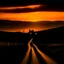 Placeholder: dark night, orange sunset colors in the sky, a lonely cottage with the lights on in the distance on a mountain, a lonely dark figure walking down the road