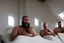 Placeholder: close up photography of two arab prisoners in cell sitting on the edge of a bed talking, muscular hairy chubby 45 years old,long beard, low lights, misery and poverty, in undershirt and boxers, little light enters from a skylight above, lights from above, photorealistic, ambient occlusion, front view from below