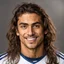 Placeholder: 85mm DSLR color photography of a very detailed headshot fitting all of head and hair in frame. 23-year-old Brazilian soccer player, with and with no facial hair and has a with a small smile, grey background has a soft look on his face has smooth long hair