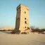 Placeholder: Fotografia di una torre moderna brutalista, che sembra . La struttura è erosa e inglobata dal paesaggio naturale, come se il tempo o l'ambiente avessero iniziato a "riassorbirla". L'atmosfera è desolata e surreale, con un ambiente circostante spoglio e sabbioso hasselblad h6d400c