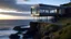 Placeholder: A modern glass and steel home perched on top of a rocky cliff overlooking the ocean at dusk