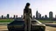 Placeholder: tall thin woman, with black straight hair, dressed in a camouflaged jumpsuit, looking out from the rear of a futuristic aircar, on a tarmac runway, with a city skyline in the distance