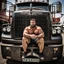 Placeholder: photography of a burly truck beefy driver at rest in truck, inside cab, shirtless, with short shorts, sweaty, massive with tattoos and short beard, Romanian, muscular, male chest, big tights, frontal view, seen from below