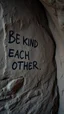 Placeholder: A hand writing On cave wall written “ BE KIND TO EACH OTHER “ an old man with bear and king hair died by the cave wall , a dim lighting great quality picture with primitive style