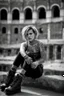 Placeholder: A portrait of a young Italian punk girl. Photo taken in front of the Colosseum. Torn T-shirt, short red skirt, heavy boots, a few tattoos and piercings, colourful teased hair voivod style, sitting pose, 135 mm lens, classic Ilford film, highly detailed skin,