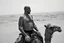 Placeholder: man looking like "Walter White" in breaking bad tv show, bald and goatee, with bare chest riding on a camel in sahara in desert, panorama, black and white photo, staring at a small town in horizon
