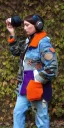 Placeholder: Brunette.thick thighs,thick calves,normal bodytype. big head. Mantle is sewed of upcycled Denim and sewed together of camouflage pieces. Pieces' color are orange, cream and purple. It is with big bright purple felt tippet and cream-colored-hood. mantle is merged with satchel. . Big AKG-style headphones (gold rings!) is merged with small felt cap with small visor. Style: Haute Couture in 1920's, N.Y.C fashion in 1996, inspired by street art. Cream latex gaiter. Her head and rest body visib