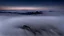 Placeholder: looking down at misty foreground and night sky background, no sun, single sharp narrow mountain peak coming through the mist in the center