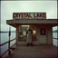 Placeholder: horror movie still, grainy color photography, faded colors, nihilistic, text "CRYSTAL LAKE MEATS" sign on a dilapidated butcher shop on a pier of a lake, foreboding, Jason Vorhees wearing a hockey mask and a butcher's apron standing in shop window, dramatic