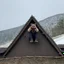 Placeholder: bearded man in a coon skin cap sitting on the roof of a rustic triangular shaped cottage holding a smart phone up over his head trying to get a signal, mountainous terrain, patchy snow on the ground and on the roof, cloudy day, photographic