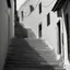 Placeholder: Calle de un pueblo de una isla italiana en verano, con escalinata, fotografía real, fotografía realizada con cámara Leica y objetivo de 50 mm, siguiendo estilo de la serie 'Ripley' emitida en Netflix, fotografía en blanco y negro, virada tonos años 50