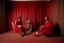 Placeholder: Un hombre vestido con traje, un hombre con pijama rojo, y una mujer vestida con traje, sentados en sillones, en una habitación de suelo de baldosas con líneas zigzagueantes, la habitación la recorre una gran cortina roja, fotografía realizada con cámara Leica y objetivo de 50 mm