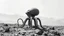 Placeholder: This is a black and white photograph featuring a surreal, abstract scene. The central subject is an octopus-like creature with an egg-shaped body, resembling a smooth, rounded shell. It has eight long, spindly legs, each ending in a cluster of smaller, bulbous appendages. The texture of the creature's body and legs is rough and uneven, giving it a somewhat organic yet alien appearance. The background is a barren, rocky landscape with faint, misty hills or mountains in the distance, creating a se