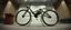 Placeholder: pinhole cam, airbrush 3d rendering, on tv screen:A five-second cinematic single take in a clean industrial presentation hall. A bizarre mechanically plausible tracked bicycle machine stands beside a presentation podium, designed to look like a giant mechanical praying mantis. Two large bicycle wheels form its main body and four thin articulated mechanical legs support it. Two long folded mechanical front arms resemble mantis forelegs