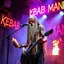 Placeholder: a very thin swedish viking man, evil with very long blonde hair (2 meter), (fat glowing eyes), he is called "KEBAB MAN" performing at death metal club, his name is "KEBAB MAN" kebab rolls On stage, kebab Neon lights and smog, and "KEBAB MAN" big sign on the wall, kebab tribal tattooes, spinning kebab machines, flames and smoke, chaos, he using gasolin and pour it on the guitar on the stage