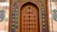 Placeholder: Middle Angle Of A Beautiful Intricately Crafted Door Of A Traditionally Designed Mosque, The Door Is Of Brown In Colors With Symmetrical Golden Floral Patterns And With Ruby Stones Symmetrically Placed On Both Sides, With Symmetrically Crafted Navy-Blue, Maroon And Floral Patterns Both Sides Of The Door With Overall Intricate Details Showing The Grandeur Of The Door.