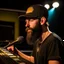 Placeholder: A short brown beard, DJ board,man with a baseball hat sing at microphone, on dancehall , microphones,, laser lights, FRONT VIEW