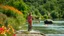 Placeholder: A lady is biking next to a river in the countryside. She moves with elegant, The background is filled with vibrant wildflowers and lush greenery along the riverbank. The camera smoothly follows her , by the way young people are swimming and a lady dives from top of a big rock to river.