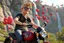 Placeholder: 3D video game characters, a short blonde haired man wearing eyeglasses, t-shirts and jeans riding a motorcycle in tuscany, hearts, waterfall, happiness