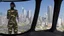 Placeholder: Tall thin woman, with straight black hair, dressed in a camouflaged jumpsuit, looking out from the rear of a futuristic aircar, on a tarmac runway, with a city skyline in the distance