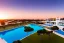 Placeholder: Sunset algarve in quinta do lago, one straight line building of 250 meters long modern luxury architecture with pool on rooftop, with green roofs and sun loungers next to pool, overlooking a tennis sport facility surrounded by pine trees, on a slope with pinus pinea, a wrap around road for low speed cars