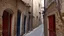 Placeholder: A narrow alley wound between limestone buildings, their walls showing centuries of wear. The eaves almost touched overhead, letting out thin cascades of water in rhythmic intervals. Low doors, some painted in bright colors like red or navy blue, contrasted with the bare facades, while small gas lamps flickered in the doorways, giving the place an air of nostalgia.