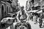 Placeholder: man looking like "Walter White" in breaking bad tv show, bald and goatee, with bare chest riding on a camel in a alley Casablanca town, panorama, black and white photo, lot of markets and people in background