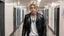 Placeholder: A young man with blond hair. He has blue eyes and is wearing an old black leather jacket, a white T-shirt, black torn jeans, and no jewelry. He is walking through the school corridor. In the background, a young woman with black hair and light gray eyes stands leaning against the school lockers. She wears a purple crystal necklace around her neck. She is wearing a white turtleneck, a navy blue skirt and black high boots. There are many students in the background wearing casual clothes.