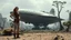 Placeholder: A skinny 50-year-old woman standing, hands on hips, in brown shorts, a shirt, and boots, standing next to a large, sleek cargo spaceship, sitting in a clearing, in a derelict alien city with trees and fungi in clumps.
