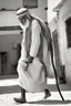 Placeholder: An old man wearing an Arabic keffiyeh, his back bent, walking barefoot, holding his cane upside down, looking back and holding his shoe in his hand.