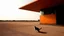 Placeholder: On the deserted runways of an abandoned airfield, the house stands quietly. The weathered orange base reflects the history of the site, while the sharp-glassed upper floor rises like a futuristic command tower. A stray cat wanders through the old hangars, pausing under the angular shadow of the building, while a photographer captures the scene from afar, intrigued by the blend of modernity and desolation.