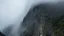 Placeholder: a massive sheer mountain cliff scaling vertically into the sky, partially obscured by dense clouds and mist. the borders — top, bottom, left, and right — fade smoothly into thick fog, while the center reveals the steep, rocky cliff face with fine texture and detail. atmospheric lighting, cinematic composition, natural colors, high contrast between fog and stone.