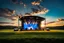 Placeholder: a big open disko stage in country side environment ,green field , at distance,blue sky pretty clouds ,sunset ,golden hour.