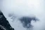 Placeholder: a massive sheer snowy mountain cliff with very sparse vegetation scaling vertically into the sky, partially obscured by dense clouds(color d0d1d5) and mist. the borders — top, bottom, left, and right — fade smoothly into thick fog, while the center reveals the steep, rocky cliff face with fine texture and detail. atmospheric lighting, cinematic composition, natural colors, high contrast between fog and stone. photography
