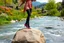 Placeholder: country side over high heels in spring wind slowly moves color full flowers,clean water fall and narrow river with waves and pretty rocks in floor ,some trees and houses ,and mountains at distant,a very beautiful Turkish lady wearing tight pants and blouse salsa dancing on a flat big rock in river , camera shots her full body